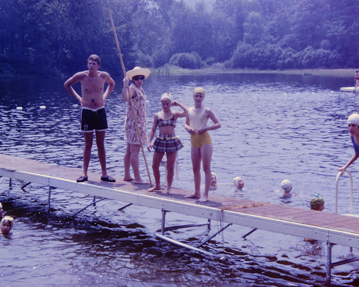 Children stand on a wooden dock extending into a lake, wearing swimsuits while others swim nearby.