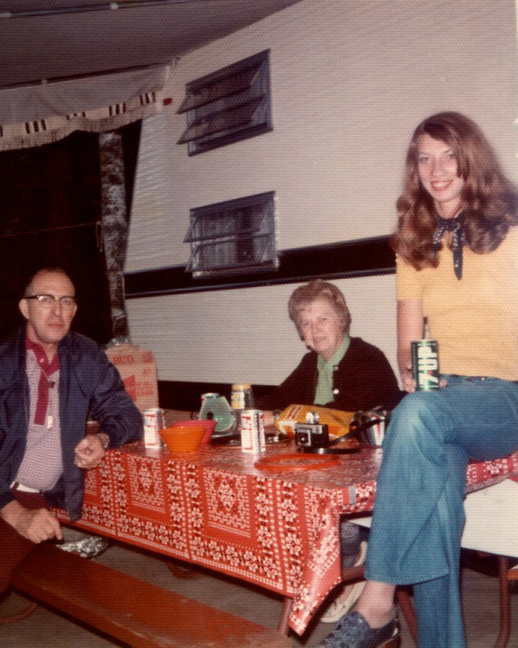 Three people at a retro indoor gathering: a man with glasses on the left, an older woman at the center, and a young woman in a yellow top on the right, seated/standing by a table with snacks and beer cans.