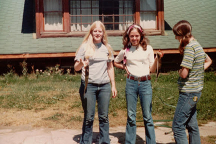 Three girls pose outside a house with a green roof; two face the camera and smile, while the girl on the right looks down holding a rope.