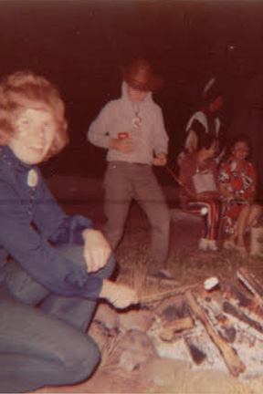 Group of people gathered around a campfire at night, with a woman in the foreground and children seated nearby, hazy vintage photo.