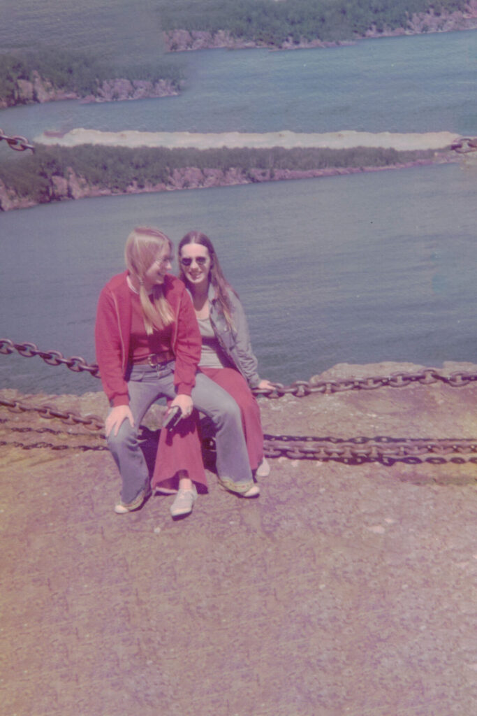 Two young women sit on a rocky ledge beside a chained barrier, smiling at the lake and each other.