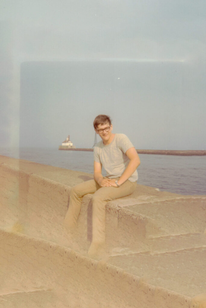 Man wearing glasses sits on a concrete seawall by the water, with a small lighthouse on a distant pier in the background.