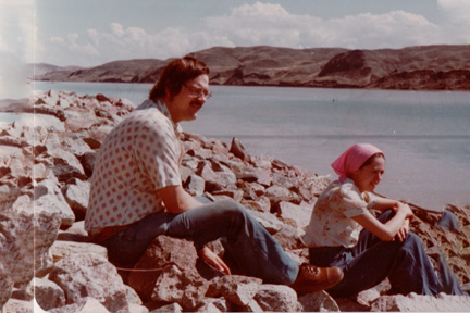 Two people sit on a rocky shoreline by a calm body of water, with hills in the distance; a man in a patterned shirt and jeans and a woman in a pink headscarf.