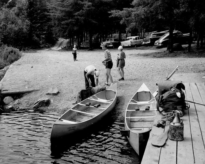 People prepare two canoes at a lakefront dock; a man leans into a canoe while others stand nearby with a wooded shoreline and parked cars in the background.