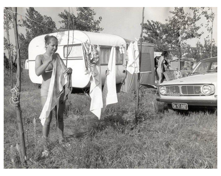 Two women in swimsuits hang laundry on a clothesline beside a camper trailer and a vintage car at a grassy campsite.