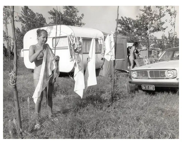 Two women in swimsuits hang laundry on a clothesline beside a camper trailer and a vintage car at a grassy campsite.
