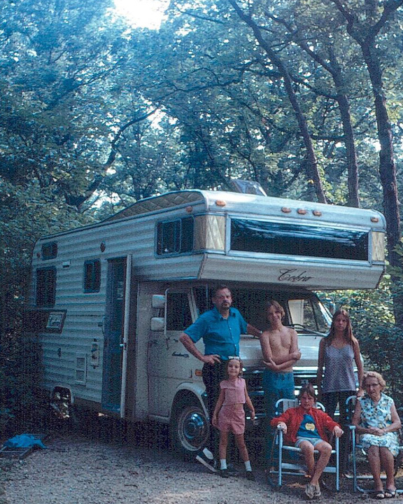 Family posing in front of a parked camper in a wooded campsite.