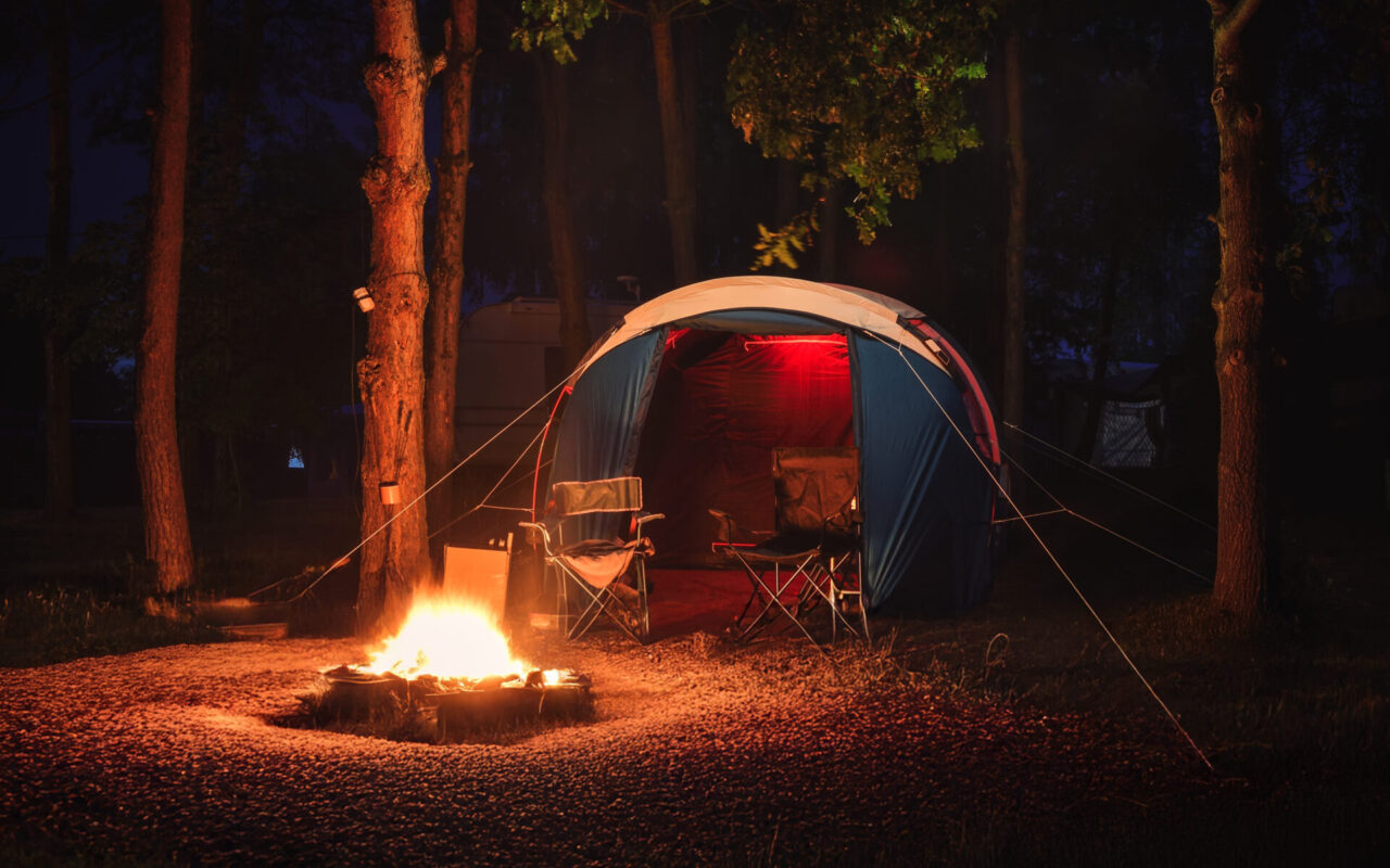 Night campsite with a blue tent, a glowing fire pit in a ring of rocks, and two folding chairs nearby under trees at night.