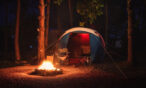 Night campsite with a blue tent, a glowing fire pit in a ring of rocks, and two folding chairs nearby under trees at night.