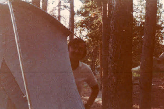 A person stands beside a large beige camping tent in a forest with tall trees and sunlight filtering through the branches.