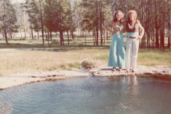 Two women standing on a rocky bank beside a small pool in a wooded park, posing for the photo.