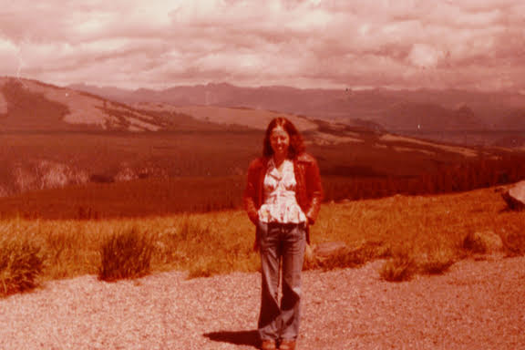 Woman standing in a dry grassy field with rolling hills and mountains in the distance, under a cloudy sky with a reddish color cast.