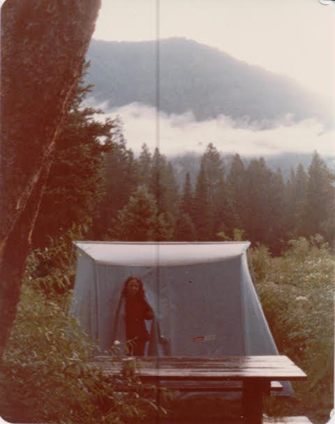 Person standing inside a white camping tent in a forest clearing, with a wooden picnic table in the foreground and mountains in the background.