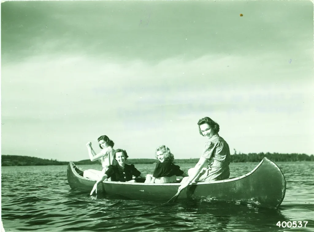 Four women sit in a canoe on a calm lake, smiling at the camera with a distant shoreline in view.