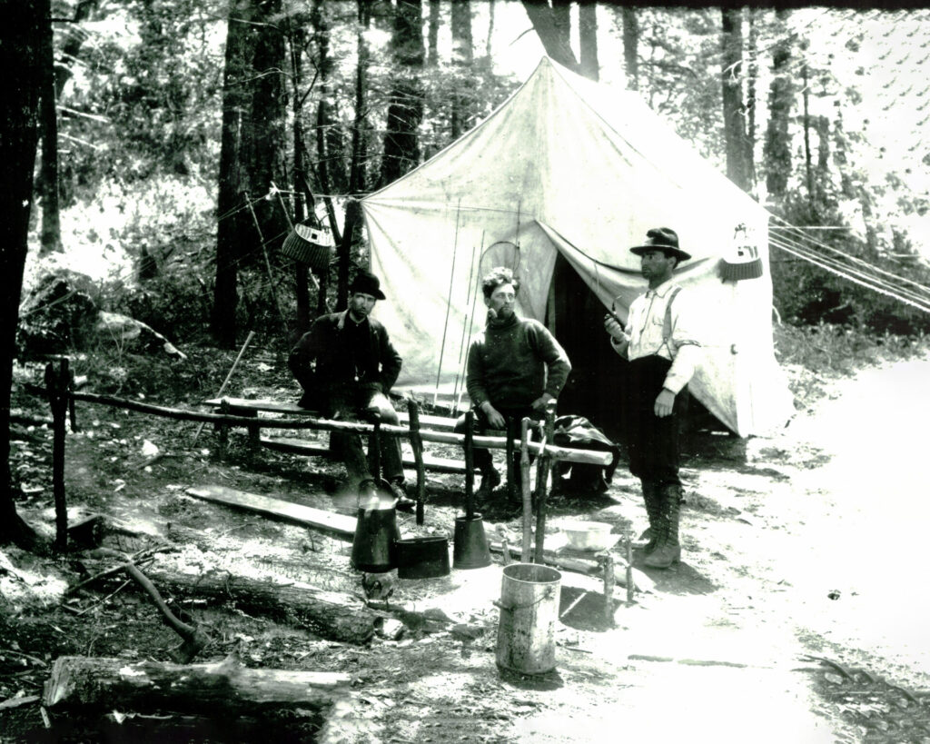 Three men sit and stand around a camp kitchen in a forest, with a canvas tent behind them and cooking pots over a fire.