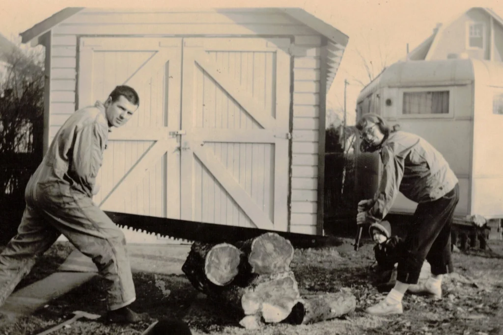 Two people and a child outdoors use a large two-person saw to cut a log into chunks, with a wooden shed and a camper in the background.