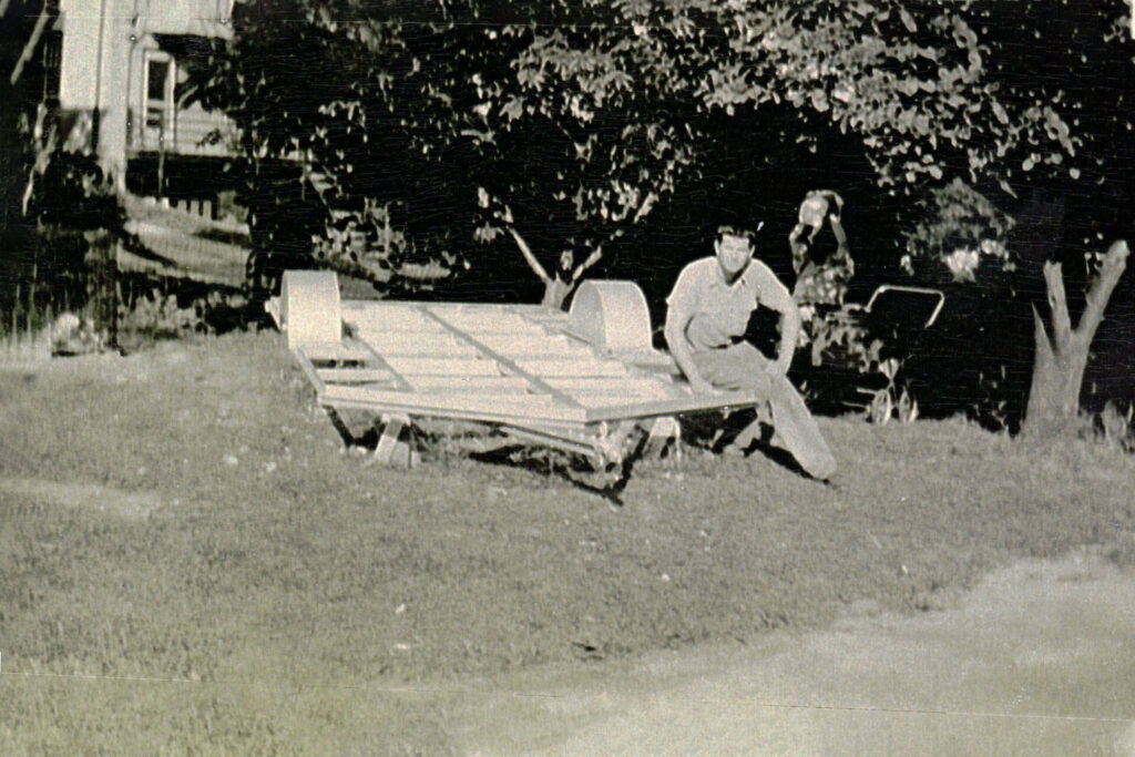 Man sits beside a damaged wooden cart on a grassy yard with trees in the background.