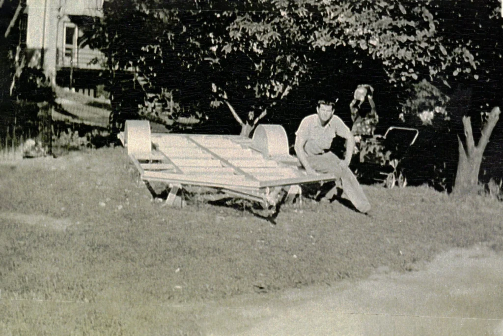 Man in a light shirt sits on a toppled wooden cart in a yard, with trees and a trailer visible in the background.
