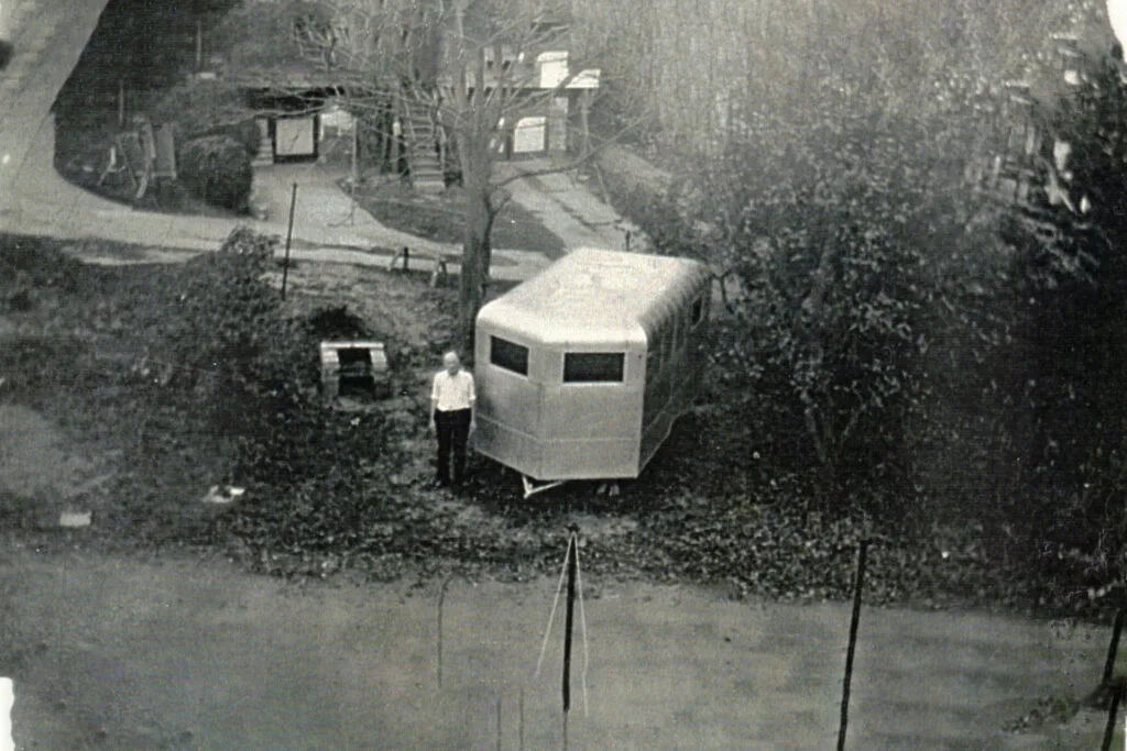 Small hexagonal white shed on a grassy slope with a person standing beside it.