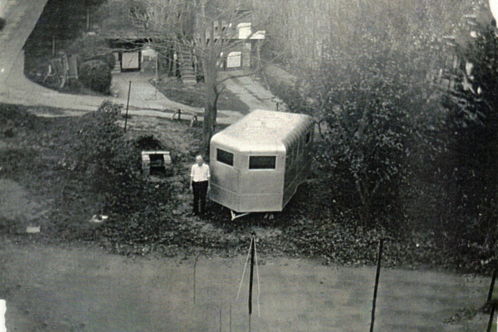 Small hexagonal playhouse with two rectangular windows in a grassy backyard; a person stands nearby.