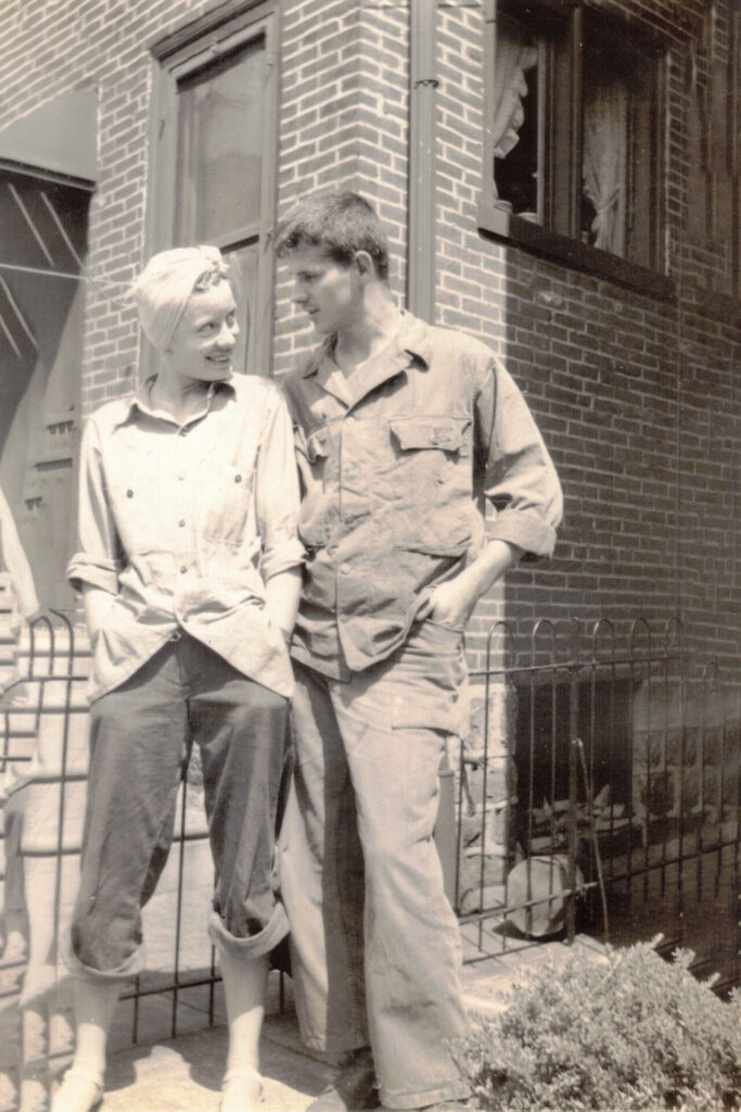 Two people in work overalls stand together outside a brick building, smiling and leaning against a small fence.