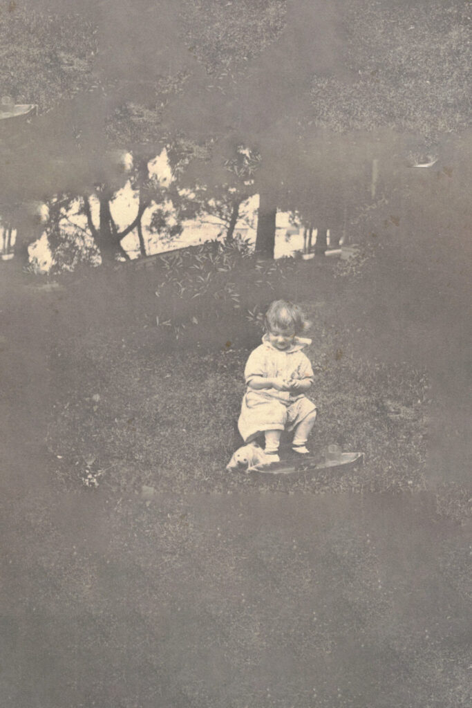 Child in a light outfit sits on a small skateboard on grass in a park, looking down at something in their hands (black-and-white photo).