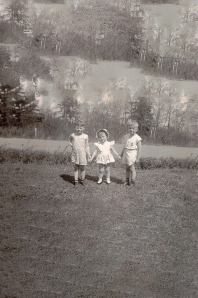 Three young children stand in a grassy yard holding hands, wearing light summer outfits in a vintage photo.