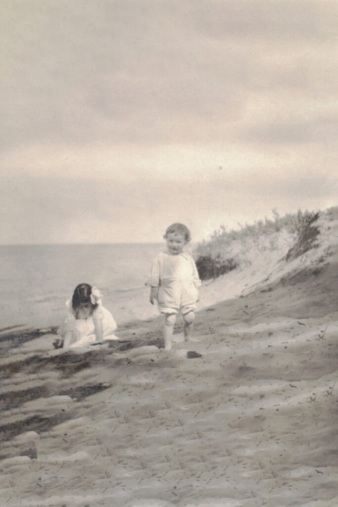 Two young children on a sandy beach near low dunes, one standing in a light outfit while the other sits near the water in the background (vintage photo).