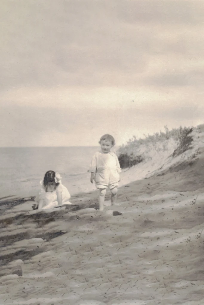 Two young children on a sandy beach near low dunes, one standing in a light outfit while the other sits near the water in the background (vintage photo).