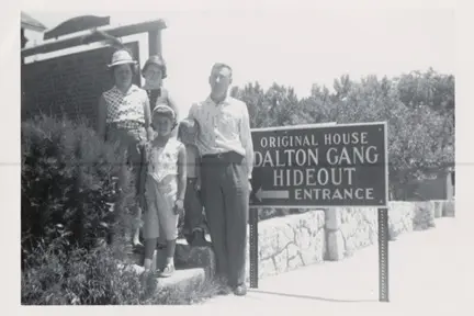 Black-and-white photo of a family of four posing beside a sign reading: Original House Dalton Gang Hideout Entrance, with a stone wall and trees in the background.