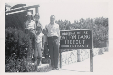 Family posing beside the Original House Dalton Gang Hideout Entrance sign at a roadside site with a stone wall behind them.