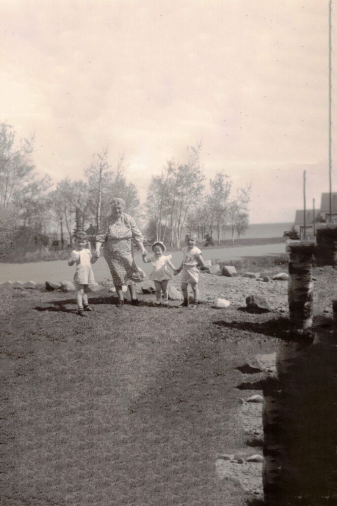 Grandmother in a patterned dress walks with three young children, holding their hands as they stroll in a grassy yard by the lake.