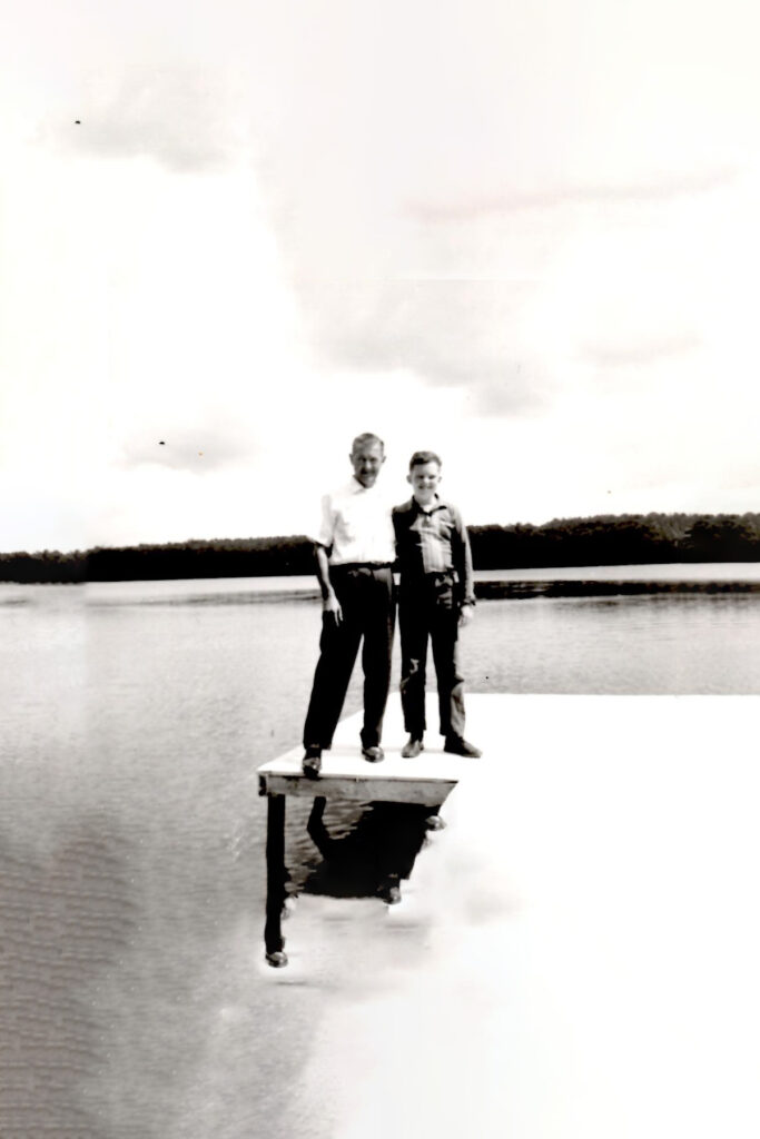 Two men stand on a small wooden dock extending into a calm lake, their reflections visible in the water.