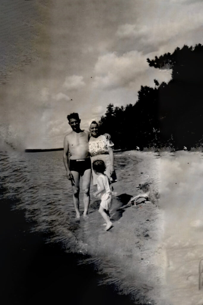 Family of three at a lake shore: man in swim trunks, woman in a patterned dress, and a small child in a swimsuit standing at the water's edge.