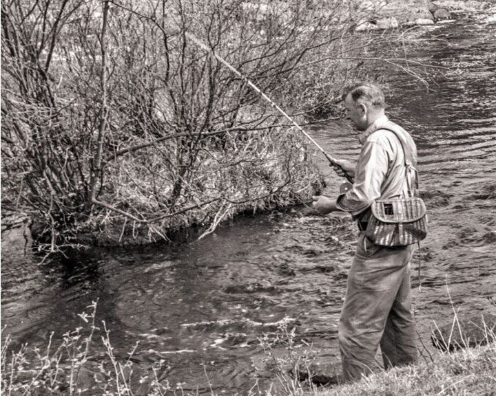 Man fishing in a river, standing on the bank with a rod and tackle vest in a black-and-white scene