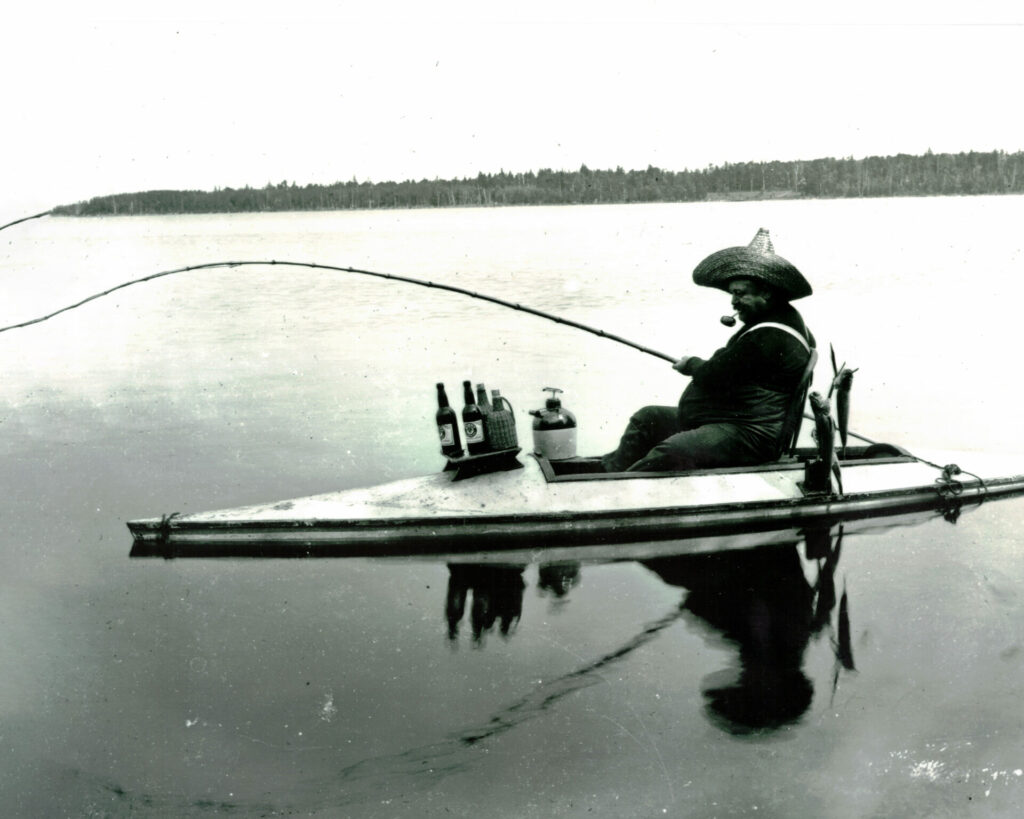 Man in a small boat fishing on calm water, wearing a wide hat and smoking a pipe, with bottles and gear aboard.