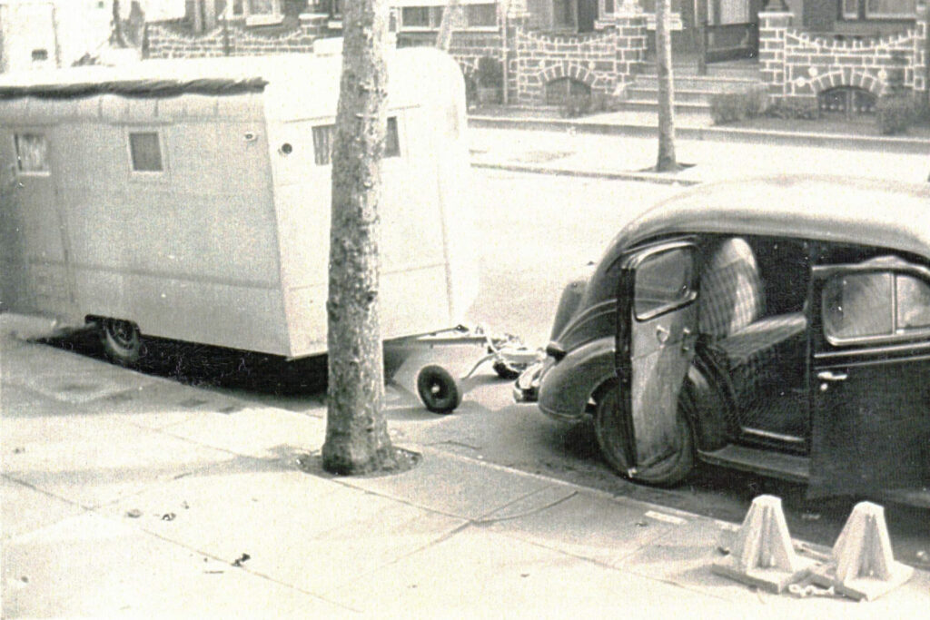 Black-and-white street scene: an old car with its front door open, parked beside a large trailer, with a tree in the foreground and brick houses in the background.