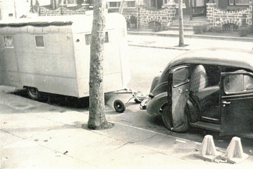Vintage car parked on a residential street with its front door open and a large trailer behind it; a tree and brick houses in the background.