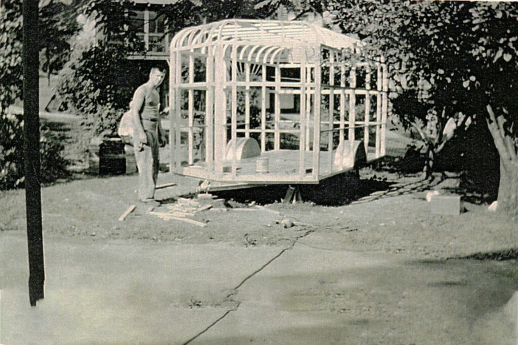 Man and woman maneuver a large wooden-framed cage on a wheeled base across a yard or lawn in front of a house trees behind.