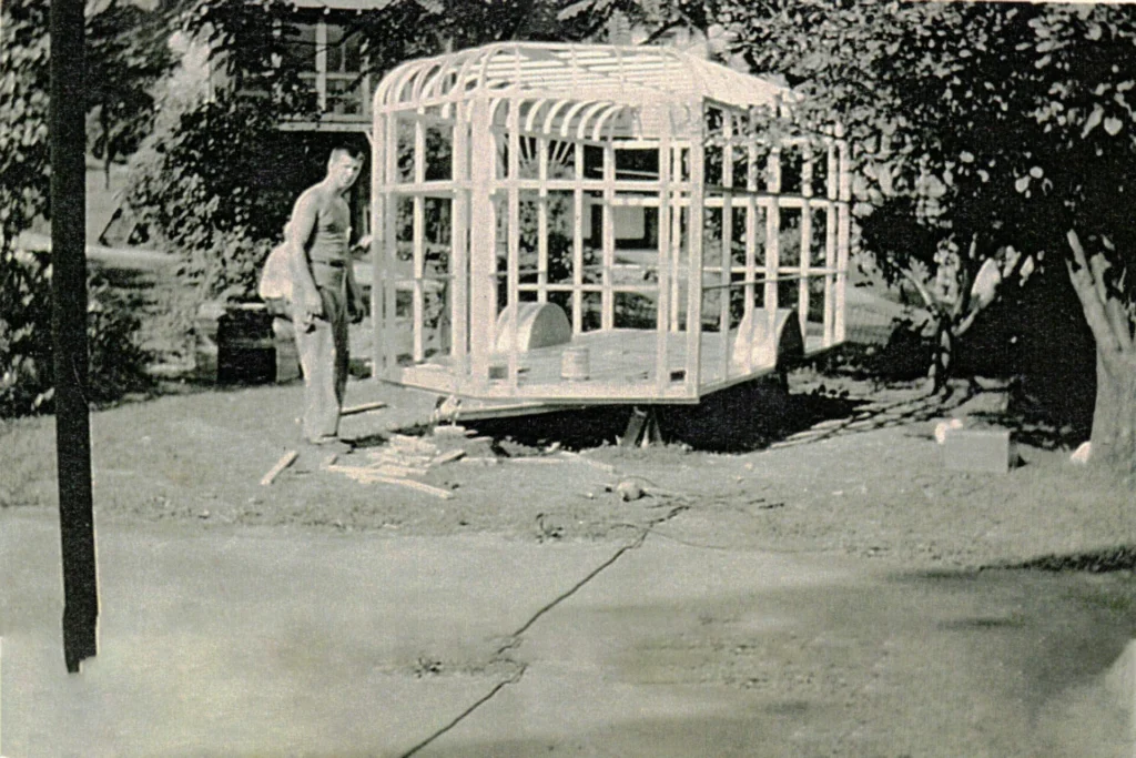 Two men stand beside a large, wheel-mounted wooden cage-like structure in a yard, as if building or moving it.