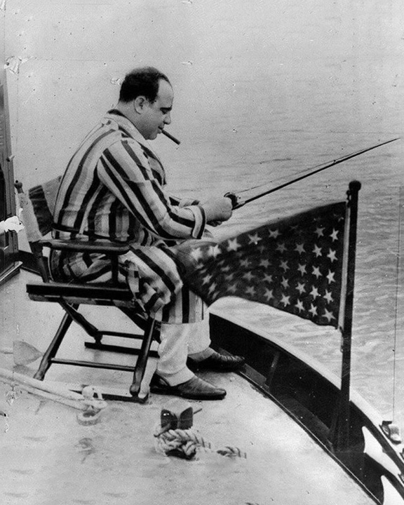 Man in a striped jumpsuit sits on a folding chair on a boat, fishing with a rod and cigarette, beside an American flag on the railing.