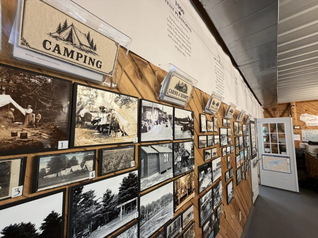 Wall of vintage black-and-white photos and display cards on a wooden wall, labeled with 'CAMPING' and 'CABINS & RESORTS' in a museum exhibit.