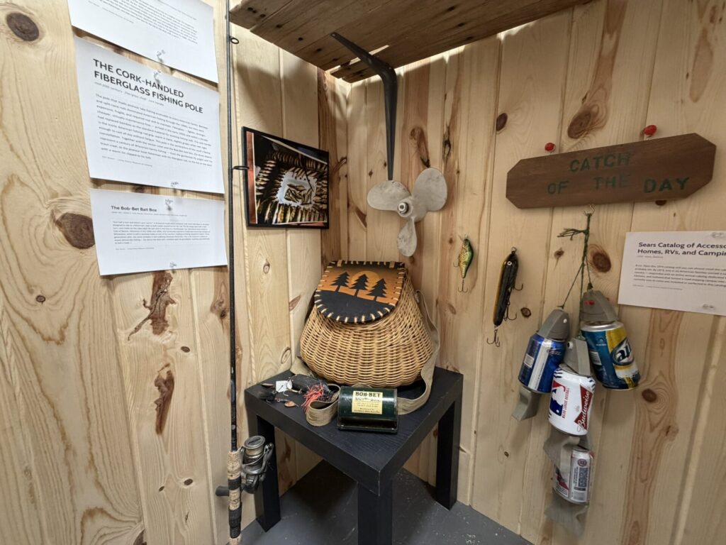 Corner display of vintage fishing gear on wood paneling: cork-handled fishing rod, wicker basket, metal propeller, fishing lures, framed photo, and a 'CATCH OF THE DAY' sign.