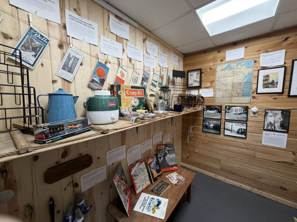 Interior of a rustic display room with vintage camping gear, maps, and posters pinned to wood-paneled walls; a wooden desk holds brochures and tools.