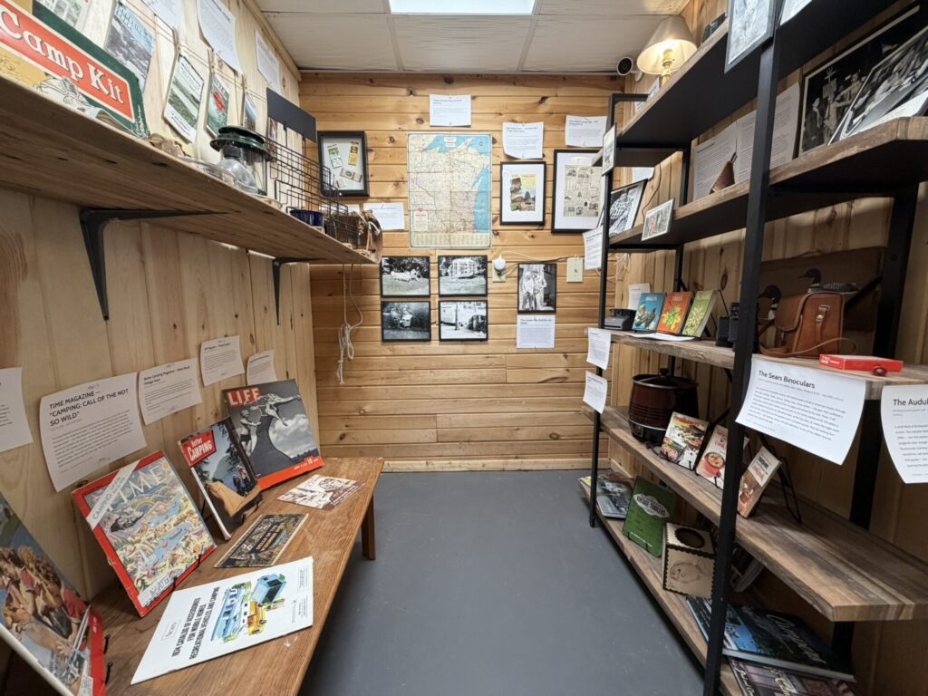 Small vintage display room with wood-panel walls, shelves and tables covered in old magazines, maps, photos and informational panels.