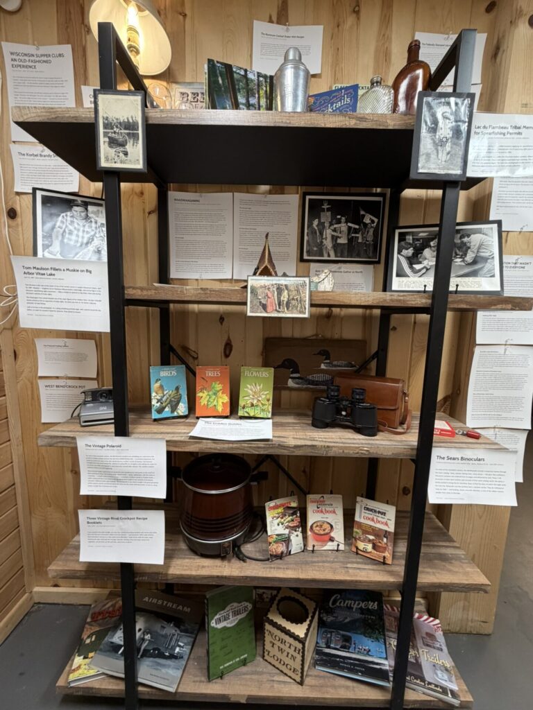 Shelf of vintage travel and cooking memorabilia with photos, documents, and books on wooden shelves used in a museum display area