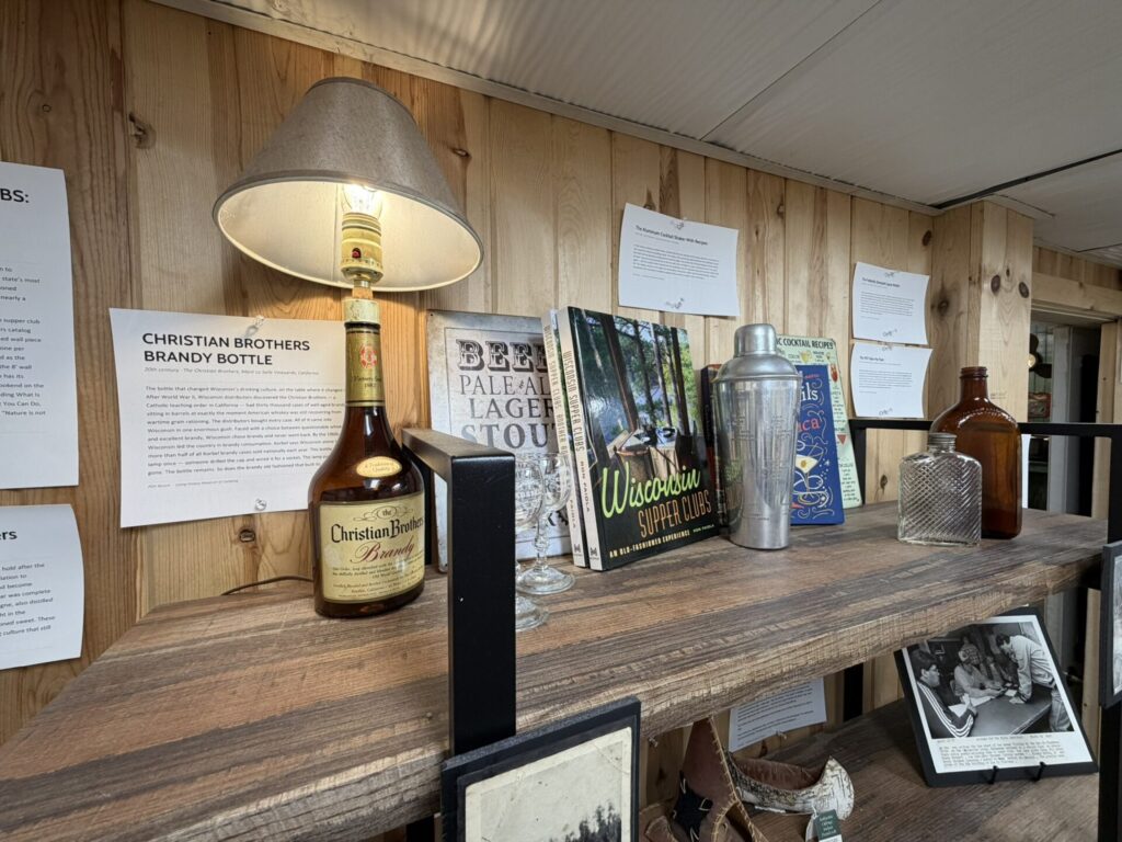 Wooden bar shelf with a Christian Brothers Brandy bottle, a lamp, metal cocktail shaker, and vintage magazines and notes on the wall.