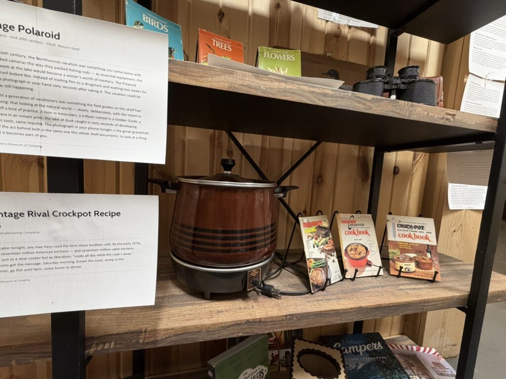 Display shelf of vintage cooking items including a Crock-Pot, old cookbooks, and informational plaques about polaroid and retro recipes for a museum exhibit.