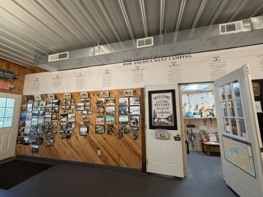 Museum exhibit with a photo collage and 'How America Went Camping' timeline on a wood-paneled wall, ceiling vents visible above