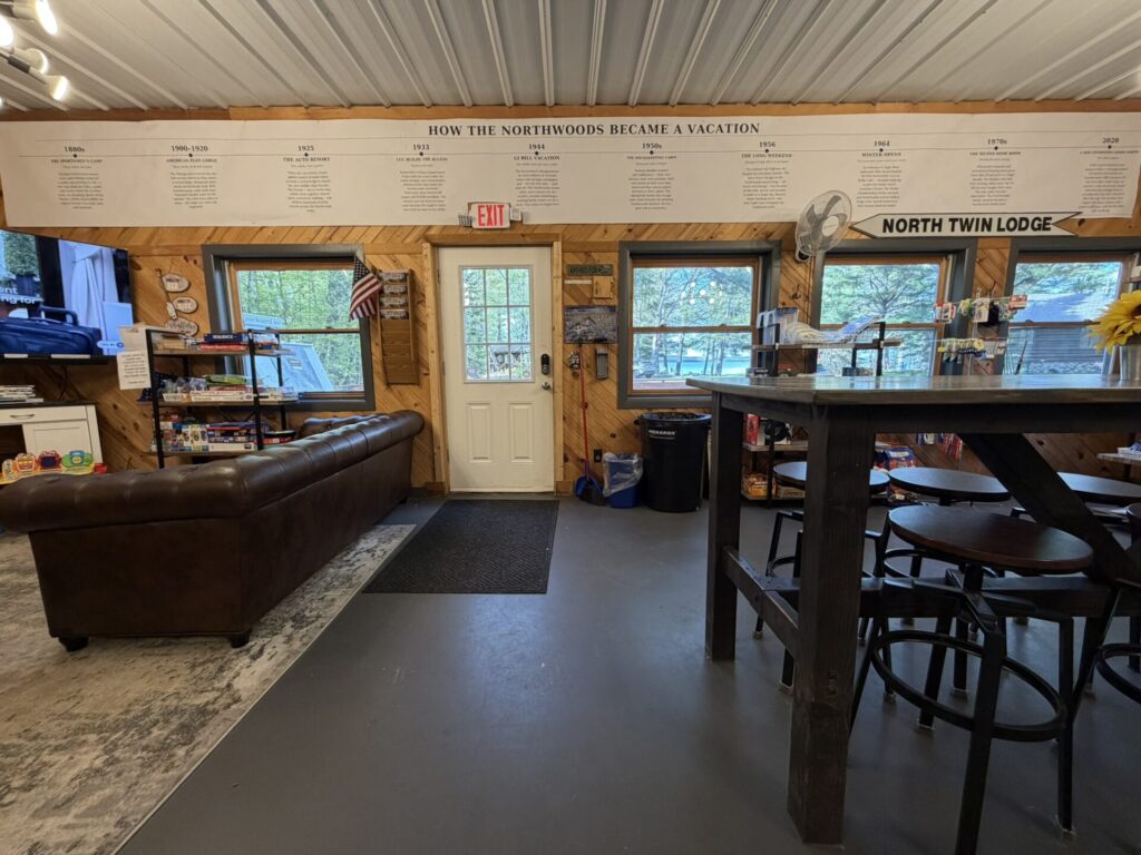 Cozy rustic lounge with a brown leather sofa, a tall wooden bar table and stools on the right, exit door center, windows to the outdoors, and a long wall timeline overhead.